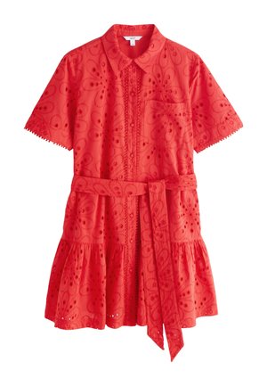 Jonge vrouw met donker haar tot op de schouders, die een felrode geborduurde blouse en een delicate parelketting draagt, kijkt recht vooruit.