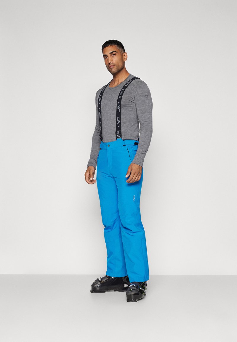 Pantalons de ski bleus avec bretelles ajustables, associés à une chemise grise à manches longues. Les pantalons présentent un design élégant et des poches pratiques.