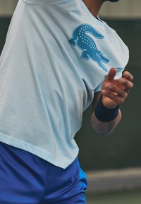 Light blue short-sleeve athletic shirt with a large blue crocodile logo on the left chest, worn with dark blue shorts, featuring a smooth texture.