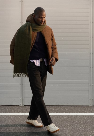 Braune Steppjacke, marineblauer Pullover, gestreiftes Hemd, schwarze Jeans, grüner Schal und weiße Sneakers mit braunen Sohlen vor einem grauen Hintergrund.