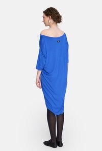 Niebieska sukienka typu off-shoulder wykonana z gładkiego materiału, z rękawami o długości trzech ćwierć i drapowaną formą, z czarną metką z tyłu.