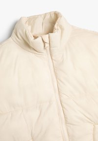 Hellbeige Pufferjacke mit hohem Kragen, Frontreißverschluss und gestepptem Design. Das Material ist glatt mit einem glänzenden Finish.