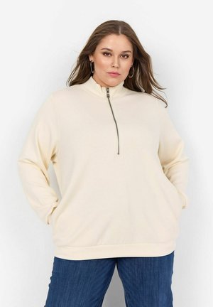 Cremefarbener Pullover mit hohem Kragen, halbem Reißverschluss vorne, Seiten Taschen und lockerer Passform. Getragen mit blauen Jeans und einer glatten Stofftextur.