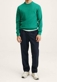 Mann in einem grünen gerippten Pullover, dunkeltblauen Hosen, weißem Unterhemd und beigen Sneakers, steht vor einem einfarbigen hellen Hintergrund.