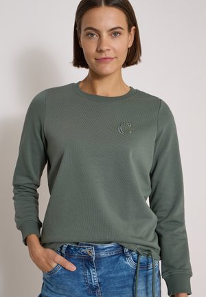 Donna con capelli corti castani che indossa una felpa verde a maniche lunghe e jeans blu con una mano in tasca su uno sfondo semplice.