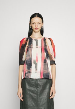 Blouse colorée avec un motif abstrait audacieux en rose, noir et crème, avec des manches transparentes et un col rond. Portée avec une jupe en cuir noir.