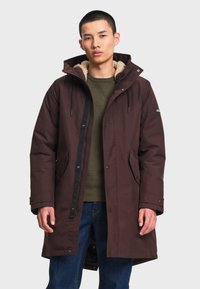 Jeune homme portant un long manteau d'hiver marron à capuche avec doublure en polaire, chemise verte et jean bleu, debout devant un fond uni.