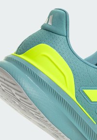 Zapatilla deportiva en color teal con parte superior de malla texturizada, acentos amarillos vibrantes, suela de goma y un collar acolchado. Presenta un diseño angular distintivo.