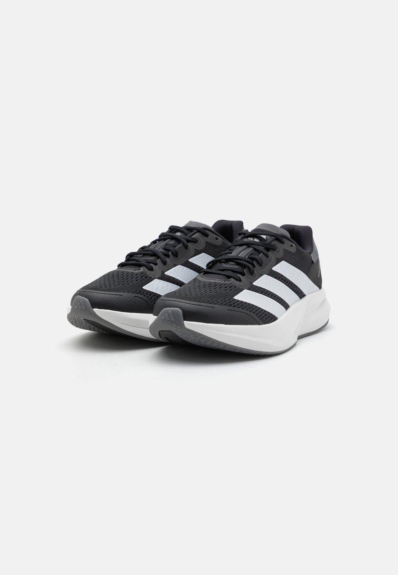 Zapatillas deportivas negras con parte superior de malla, con tres rayas blancas, lengüeta acolchada y suela blanca. Incluye detalles texturizados en el talón.