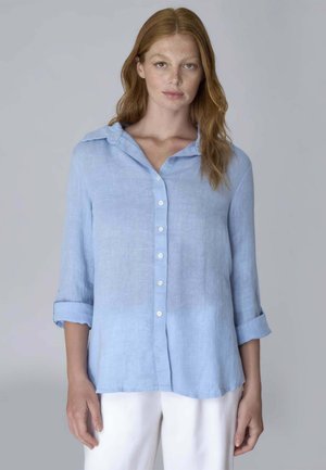 Donna con lunghi capelli rossi, che indossa una camicia azzurro chiaro abbottonata con le maniche rimboccate e pantaloni bianchi, in piedi davanti a uno sfondo grigio neutro.