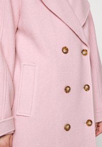 Hellrosa Wollmantel mit doppelreihigem Design, großen braunen Knöpfen, Spitzkragen und Seitentasche. Strukturierter Stoff mit weicher Oberfläche.