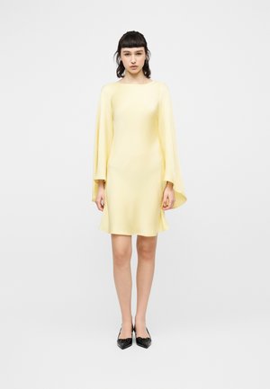 Kvinde står med fronten mod kameraet iført en lys gul knælængde kjole med lange, flydende ærmer og sorte spidse hæle på en ensfarvet baggrund.