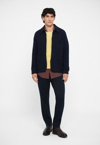 Sotilasharma villatakki, jossa on kaksi etutaskua, keltainen neuleen päällä ja ruudullinen paita pilkistämässä alhaalta, yhdistettynä tummiin housuihin.