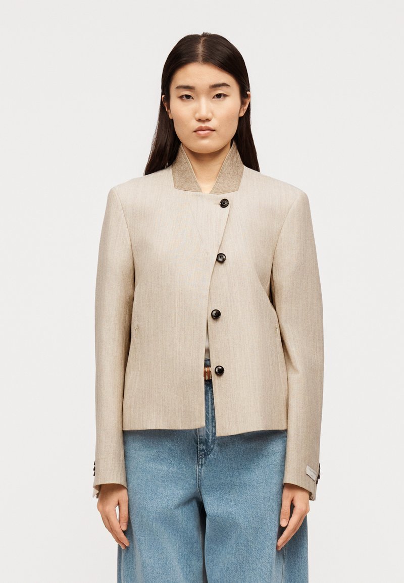 Γυναίκα με μακριά σκούρα μαλλιά, φορώντας μια μπ εζ ασύμμετρη μπουφάν με κουμπιά και ανοιχτό μπλε τζιν παντελόνι με φαρδιά ποδάρια, στέκεται μπροστά σε λευκό φόντο.