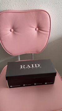 črn pravokotni kovček z logotipom "RAID LONDON" v beli barvi, postavljen na svetlo roza oblazinjenem stolu s krožnimi gumbi. Gladka tekstura.