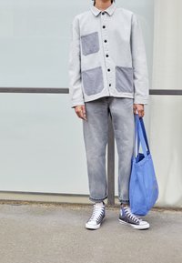 Szara kurtka dżinsowa z dwoma ciemnoszarymi naszywanymi kieszeniami, zestawiona z jasnoszarymi dżinsami i szarymi sneakerami za kostkę. Niebieska torba typu tote trzymana w ręce.