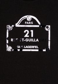 Svart t-shirt med en metallisk rektangulär grafik som har texten "PARIS 21 RUE ST-GUILLAUME" och "KARL LAGERFELD" i fet vit lettering.