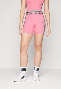 Rosa idrettsshorts laget av elastisk stoff med tettsittende passform. Har svarte detaljer og tre striper på sidene. Hvite idrettssko er iført.
