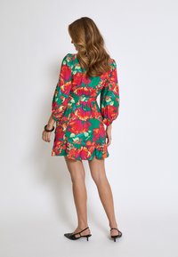 Kvetinové mini šaty so zeleným základom, s červenými a oranžovými kvetmi. Dlhé rukávy, volánkový lem a detaily s pritiahnutým pásom. Čierne slingback topánky.