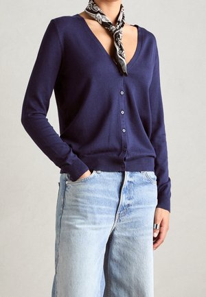 Cardigan bleu marine en tissu doux, design en V, bouton devant, manches longues, associé à un jean bleu clair et une écharpe à motifs autour du cou.
