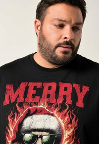 Muž s plnovousem, který má na sobě černé tričko s grafikou hořící lebky a velkým červeným nápisem "MERRY", dívá se mírně dolů a na stranu.