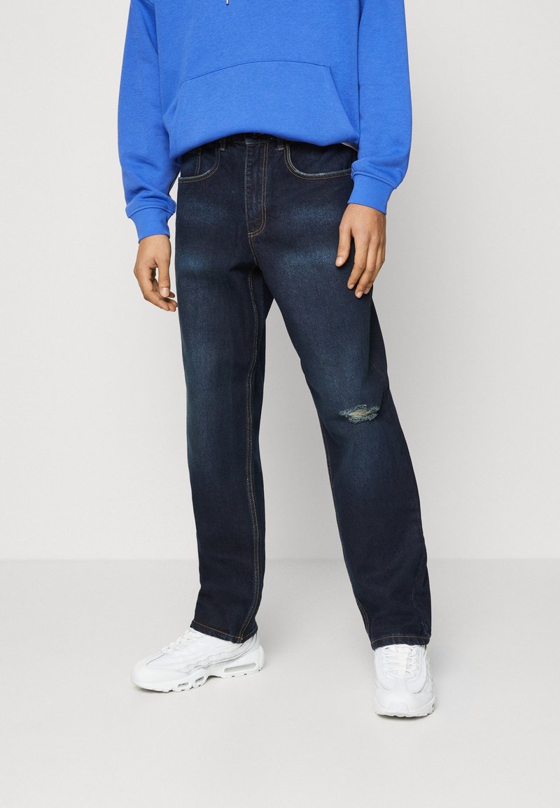 Vaqueros de mezclilla azul oscuro con un corte relajado, que presentan un desgarrón visible y costuras en contraste. Combinados con un sudadera azul y zapatillas blancas.