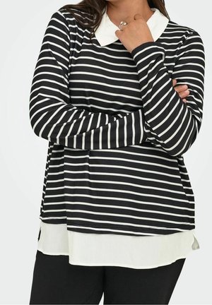 Personne portant une chemise à manches longues rayée noir et blanc avec un col blanc et un ourlet blanc superposé, les bras croisés, main près du menton.