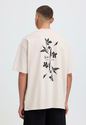 Homme portant un T-shirt beige oversize avec un motif floral noir et le texte "Solid EST. 1999" au dos, associé à un short noir.