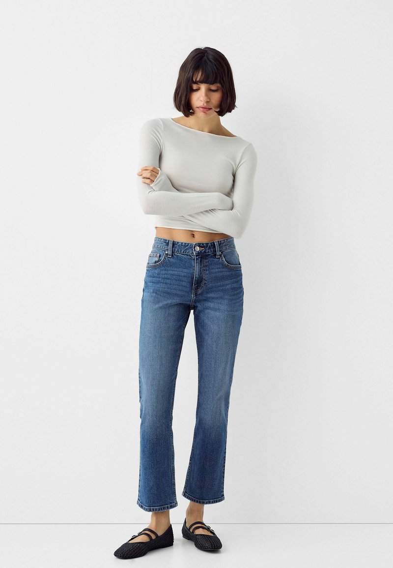 Top corto a maniche lunghe grigio chiaro, jeans azzurri a vita alta e scarpe basse nere intrecciate; design aderente con leggero elastico e orlo sfrangiato.