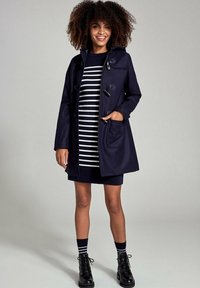 Marineblauwe jas met togglesluitingen, voorzien van een capuchon en voorzakken, gedragen over een zwart-wit gestreepte jurk. Zwarte veterschoenen en gestreepte sokken.
