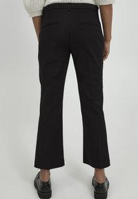 Person wearing black tailored dress pants and black shoes, standing with back facing the camera against a plain light background.