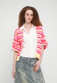 Πλεκτή ζακέτα με ρίγες σε ροζ, πορτοκαλί και κίτρινο χρώμα. Μανίκια τριών τετάρτων και ανοιχτό μπροστινό μέρος, συνδυασμένη με λευκό πουκάμισο με κουμπιά και γκρι φούστα με σχέδιο.