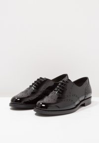 Une paire de chaussures brogues en cuir verni noir avec un design à l'aile et des lacets, présentée sur une surface blanche contre un fond clair.