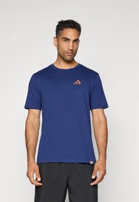 Man in een blauw T-shirt met korte mouwen met een klein oranje bergenlogo op de borst, gecombineerd met zwarte broek, staande tegen een effen achtergrond.