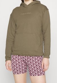 Sudadera con capucha verde oliva con un bolsillo frontal y logo bordado, combinada con pantalones cortos rojos con un patrón de diseños blanco en forma de ola.