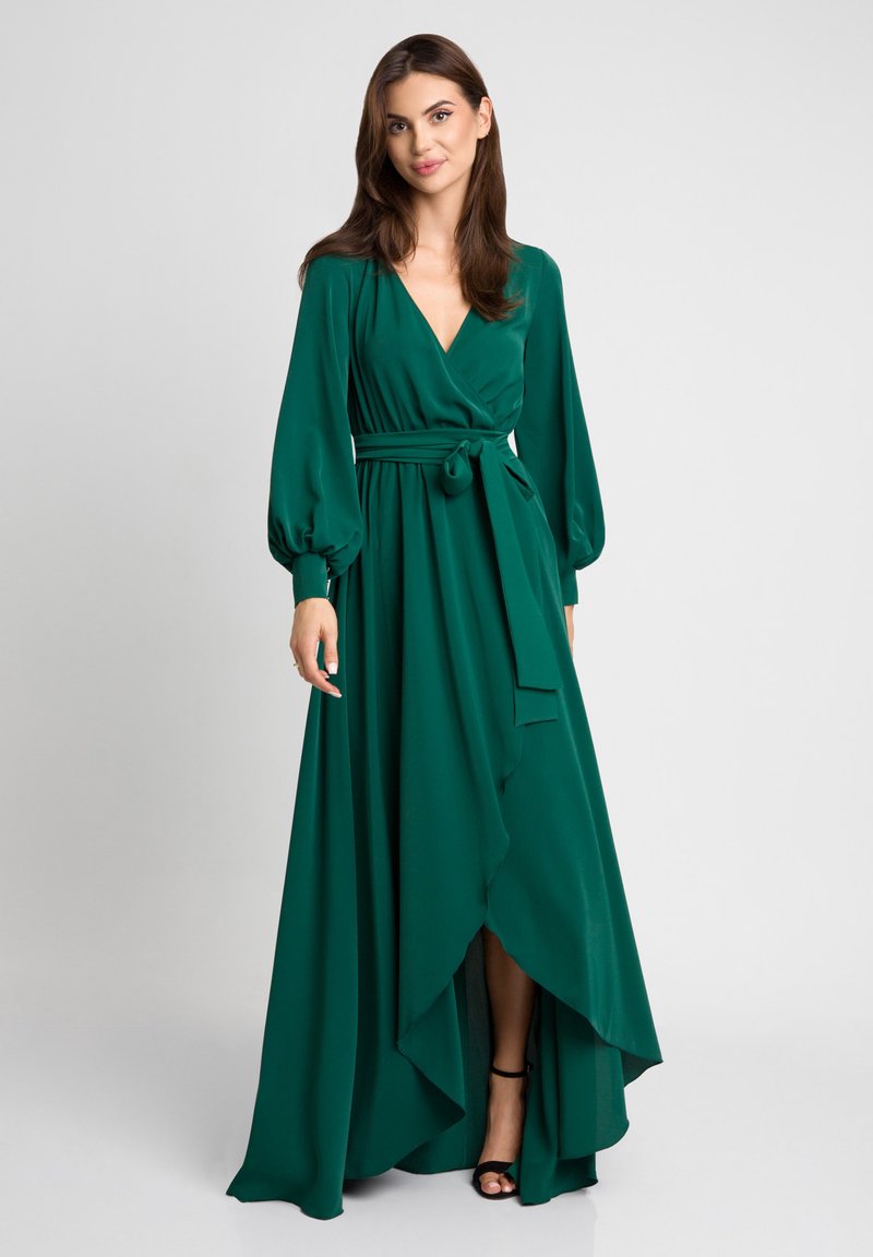 Długa zielona sukienka w gładkim materiale, z głębokim dekoltem w serek, bufiastymi rękawami i asymetrycznym dołem. Związany w pasie podkreśla sylwetkę.