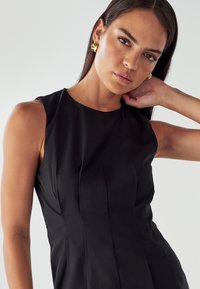 Moteris su ilgais tamsiais plaukais, vilkinti be rankovių juodą suknelę, aukso auskarais ir pozuojanti su viena ranka už kaklo prieš paprastą foną.