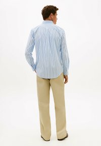 Uomo di spalle che indossa una camicia a righe verticali azzurro chiaro, pantaloni beige e scarpe marrone scuro su sfondo bianco.