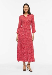 Femme souriante, portant une robe rouge à manches longues, longueur cheville, à pois blancs, avec des sandales noires à talons hauts, debout sur un fond blanc.