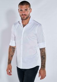 Weißes, kurzärmliges Hemd mit umgeschlagenen Ärmeln, aus einem glatten Stoff gefertigt. Verfügt über einen klassischen Kragen und einen dezenten Glanz. Getragen mit dunklen Jeans.