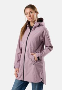 Mladá žena v fialovém nepromokavém plášti s kapucí, se zipovými kapsami a černým zipem, stojící s jednou rukou v kapse.