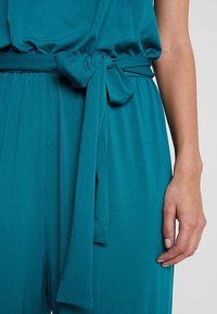 Combinaison bleu sarcelle avec une ceinture en tissu nouée à la taille et une main droite reposant à côté de la hanche sur un fond clair.