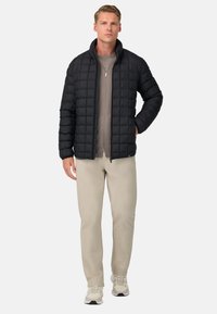 Černá prošívaná péřová bunda s vysokým límcem, nošená přes šedý top. Světle béžové kalhoty a šedé tenisky dotvářejí tento outfit.