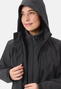 Giacca nera impermeabile con cappuccio, chiusura con zip, interno morbido e dettagli in Velcro. Logo del marchio visibile sul davanti.