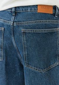 Un jean en denim bleu foncé présente une étiquette en cuir avec "BZB" au niveau de la taille, cinq poches avec des coutures contrastées et une finition texturée.