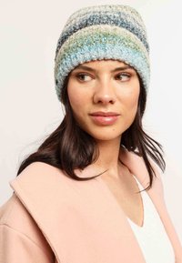 Donna con lunghi capelli scuri che indossa un berretti in maglia a strisce pastello e un cappotto rosa pallido, che guarda leggermente di lato.
