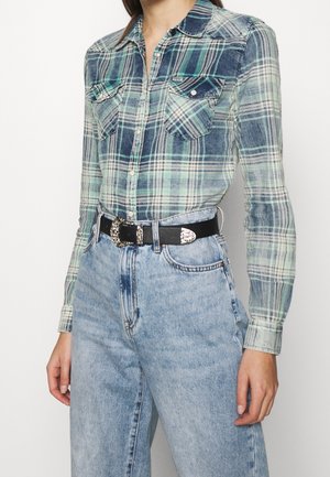 Woman wearing green and blue plaid button-up shirt tucked into high-waisted light blue jeans with a black belt with decorative silver buckle.