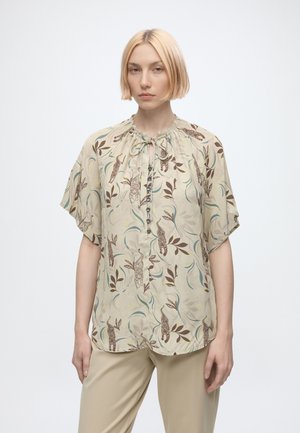 Femme aux cheveux blonds courts portant un chemisier beige avec des motifs de tigre et de feuilles et un pantalon beige, debout devant un fond blanc uni.