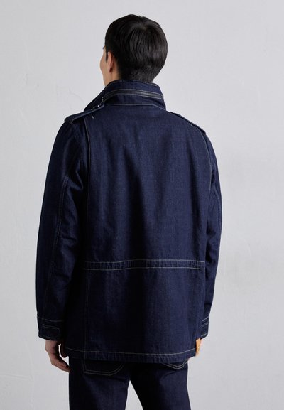 Denim jacket in dark blue with a high collar, shoulder epaulets, and contrasting yellow stitching. Visible back seams and side pockets.