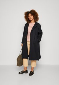 Dobbeltspent marineblå trench coat, lett stoff, vist over en rosastripete topp, sammen med kremfargede capribukser og svarte loafers.